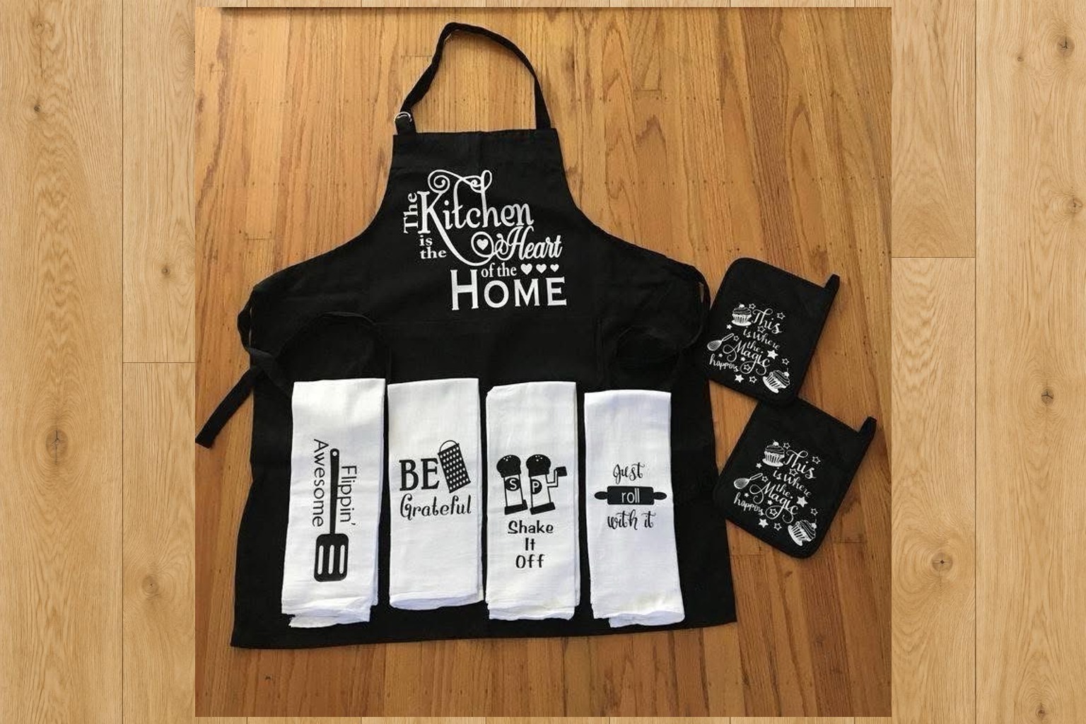A handmade kitchen set laid out on a light wood floor: a black apron printed The Kitchen is the Heart of the HOME, four white kitchen towels with sayings on them, and two black pot holders